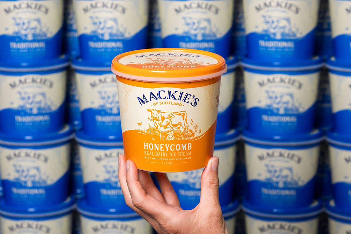 A person holds up a tub of Mackie's of Scotland Honeycomb Ice Cream with a wall of tubs of Mackie's of Scotland Traditional Ice Cream behind it.