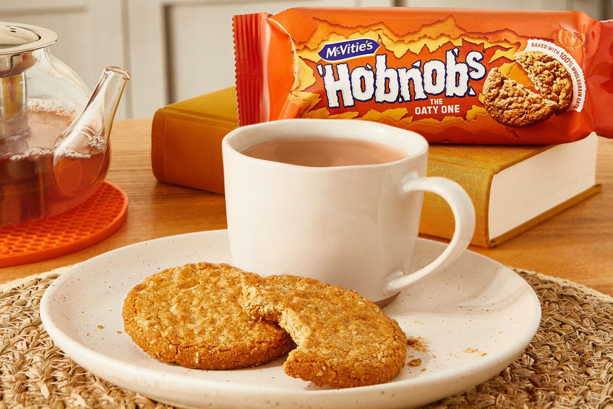 A plate with a cup of tea and HobNob biscuits on it are in the foreground with a packet of McVitie's Hobnobs in the background and a glass teapot next to the plate.