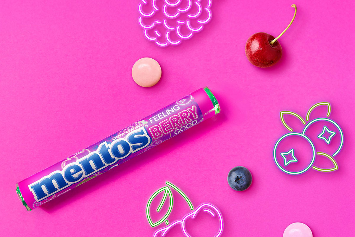 Promotional image of launch for Mentos Feeling Berry Good single pack format launch with pack of the sweets with neon signs of fruit with pieces of fruit against a pink background.