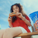 New York Bakery back on the small screen A young girl sits on a bench enjoying a New York Bakery bagel with a bag of the bagels next to her and the New York skyline in the background.