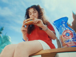 New York Bakery back on the small screen A young girl sits on a bench enjoying a New York Bakery bagel with a bag of the bagels next to her and the New York skyline in the background.