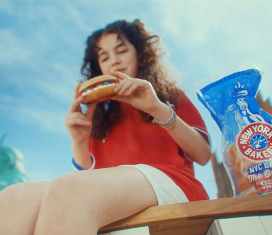 New York Bakery back on the small screen A young girl sits on a bench enjoying a New York Bakery bagel with a bag of the bagels next to her and the New York skyline in the background.