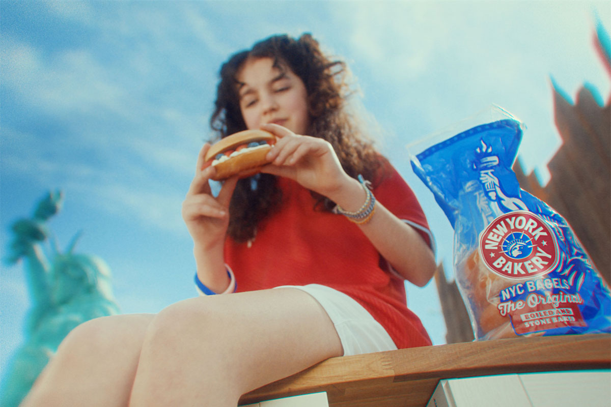 A young girl sits on a bench enjoying a New York Bakery bagel with a bag of the bagels next to her and the New York skyline in the background.
