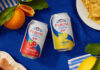 Peroni 0.0% expands alcohol free range with flavoured offer Cans of Peroni 0.0% Arancia Rossa and Limone di Sicilia sit at the middle of a table with a blue tablecloth on top with lemons and blood oranges around the cans and food.