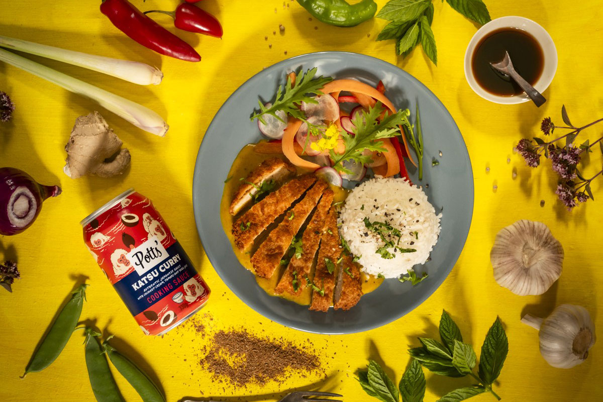 A plate of Katsu Curry is next to a Potts' can of the curry sauce with ingredients lying around the pair of them.