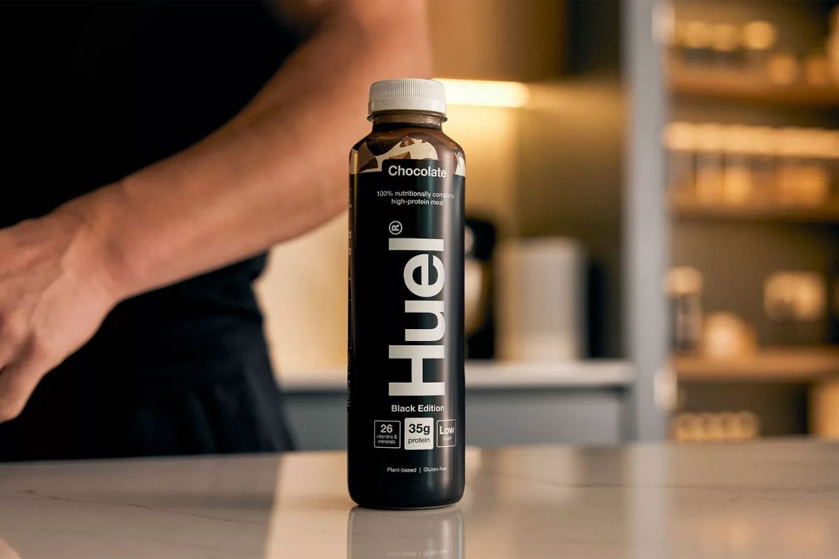 A bottle of Huel Chocolate Black Edition sits on a countertop with a person behind the bottle.