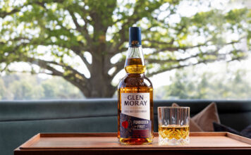 Glen Moray beckons temptation with Forbidden Fruit launch A bottle of Glen Moray Forbidden Fruit stands on a wooden tray with a rocks glass beside it. In the background is a tree.