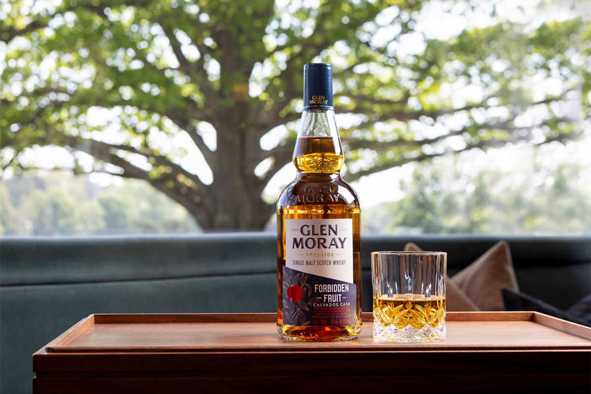 A bottle of Glen Moray Forbidden Fruit stands on a wooden tray with a rocks glass beside it. In the background is a tree.