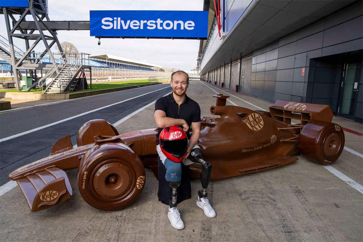 Billy Monger, former F1 driver, sits in front of a life-sized chocolate F1 car with the KitKat logo on it and the starting line of Silverstone Circuit.