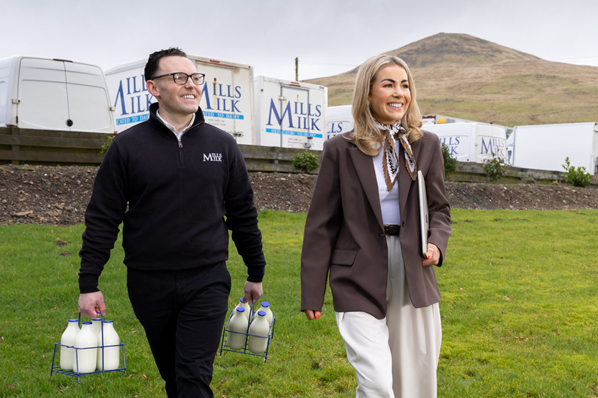 Members of the Mills Milk team stand in a field with branded vans behind them and a hill in the distance.