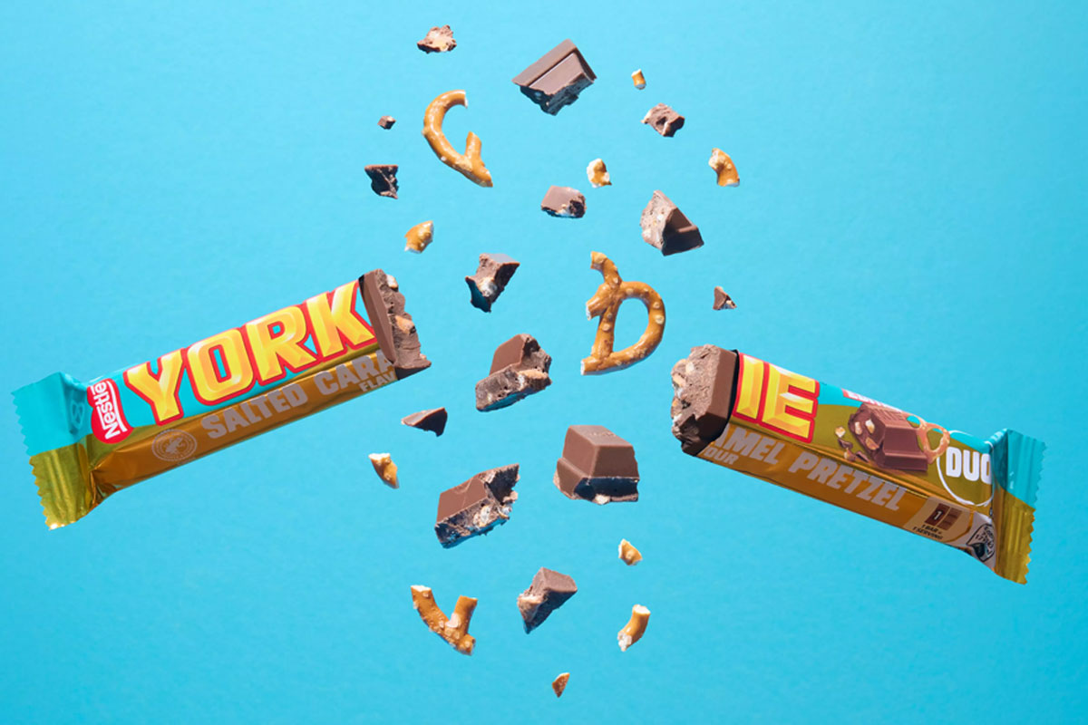 A Yorkie Salted Caramel Pretzel bar is broken in two with pieces flying out from it against a blue background.