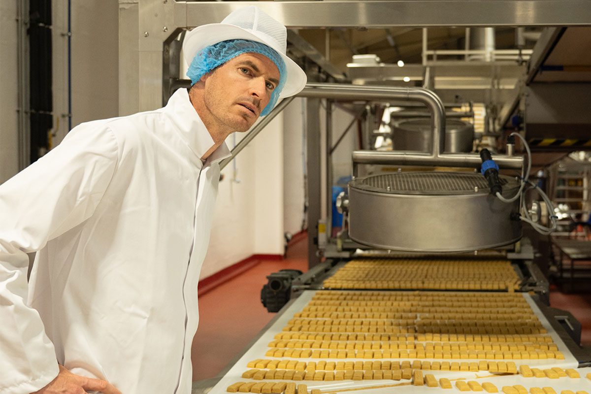 Andy Murray stands over a conveyer belt with shortbread on it.