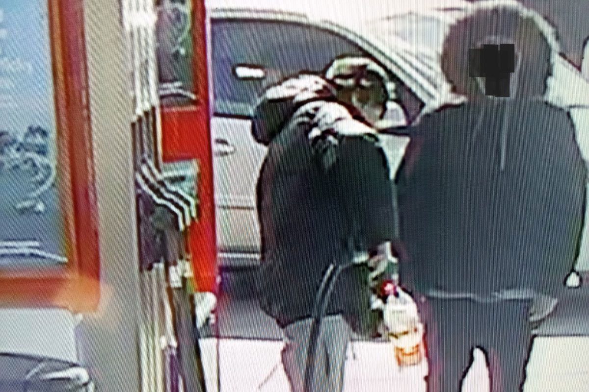 Two people stand at a forecourt fuel station as one of them fills up a plastic bottle with fuel to steal.
