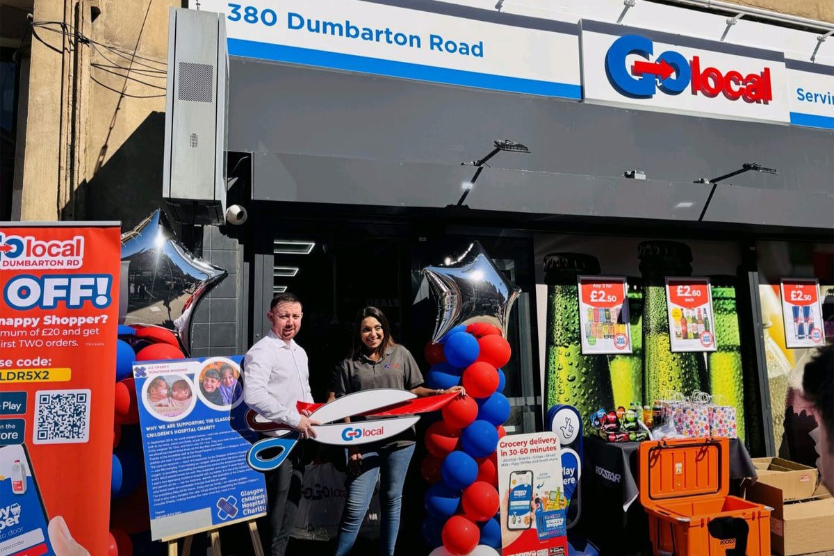 Two people stand outside Go Local Dumbarton Road holding a pair of novelty scissors at the store's door to mark the opening of the location.