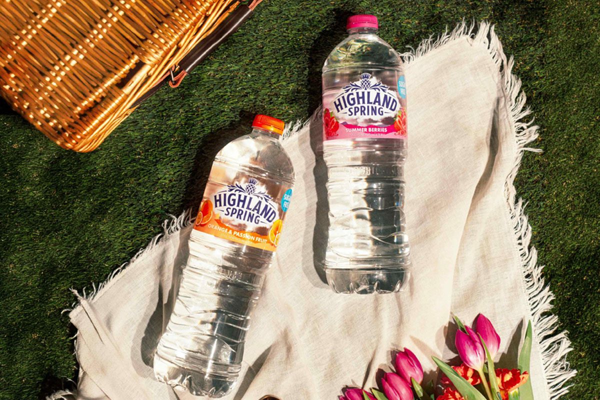 Bottles of Highland Spring Orange & Passionfruit and Summer Berries lie on a picnic scene with a wicker basket in the background.