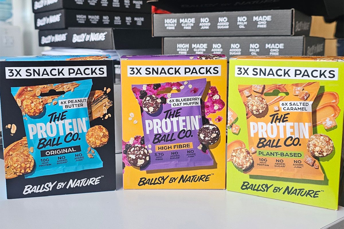 Packs of The Protein Ball Co Snack Packs stand upright on a table in Peanut Butter, Blueberry Oat Muffin and Salted Caramel flavours.