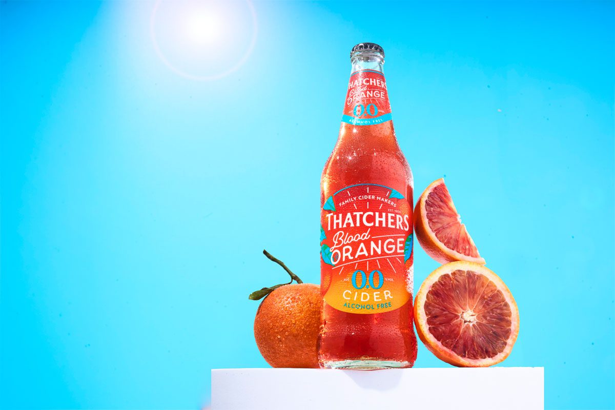 A bottle of Thatchers Blood Orange 0.0% stands on a white podium with blood oranges around it with a sunny background.