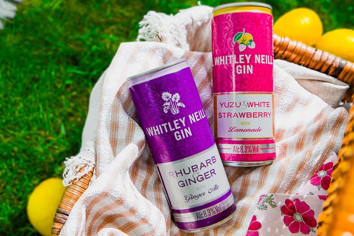 Cans of Whitley Neill Rhubarb & Ginger with Ginger Ale and Yuzu & White Strawberry with Lemonade sit in a wicker basket on grass with lemons around the basket.