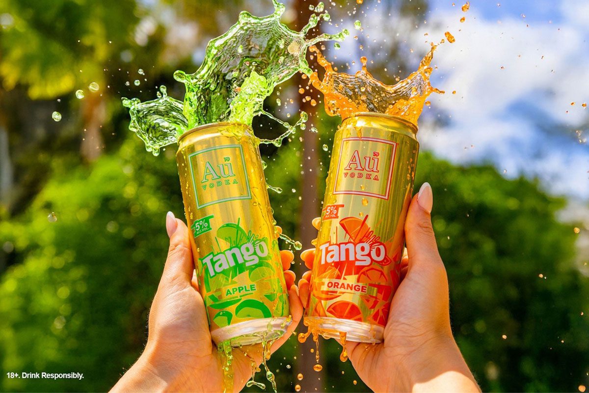 A person holds up two cans of Au Vodka Tango one in Apple and one in Orange with the drinks spilling out the top of the cans.