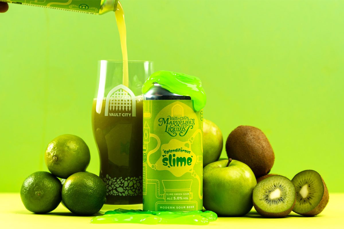 A can of Vault City Splendiferous Slime stands amongst green fruit with green slime on top of the can whilst the beer is poured out into a Vault City glass behind the can.