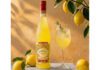 Luxardo introduces new sustainable bottle for Limoncello The 2026 redesign for Luxardo Limoncello stands on a table next to a wine glass with a cocktail serve in it and lemons have been placed around the two with a lemon tree hanging above them.