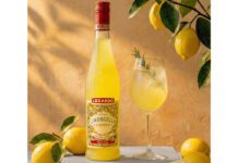 Luxardo introduces new sustainable bottle for Limoncello The 2026 redesign for Luxardo Limoncello stands on a table next to a wine glass with a cocktail serve in it and lemons have been placed around the two with a lemon tree hanging above them.