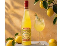 Luxardo introduces new sustainable bottle for Limoncello The 2026 redesign for Luxardo Limoncello stands on a table next to a wine glass with a cocktail serve in it and lemons have been placed around the two with a lemon tree hanging above them.
