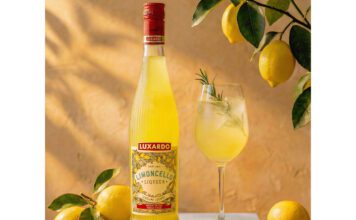 Luxardo introduces new sustainable bottle for Limoncello The 2026 redesign for Luxardo Limoncello stands on a table next to a wine glass with a cocktail serve in it and lemons have been placed around the two with a lemon tree hanging above them.