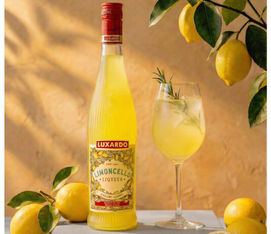 Luxardo introduces new sustainable bottle for Limoncello The 2026 redesign for Luxardo Limoncello stands on a table next to a wine glass with a cocktail serve in it and lemons have been placed around the two with a lemon tree hanging above them.