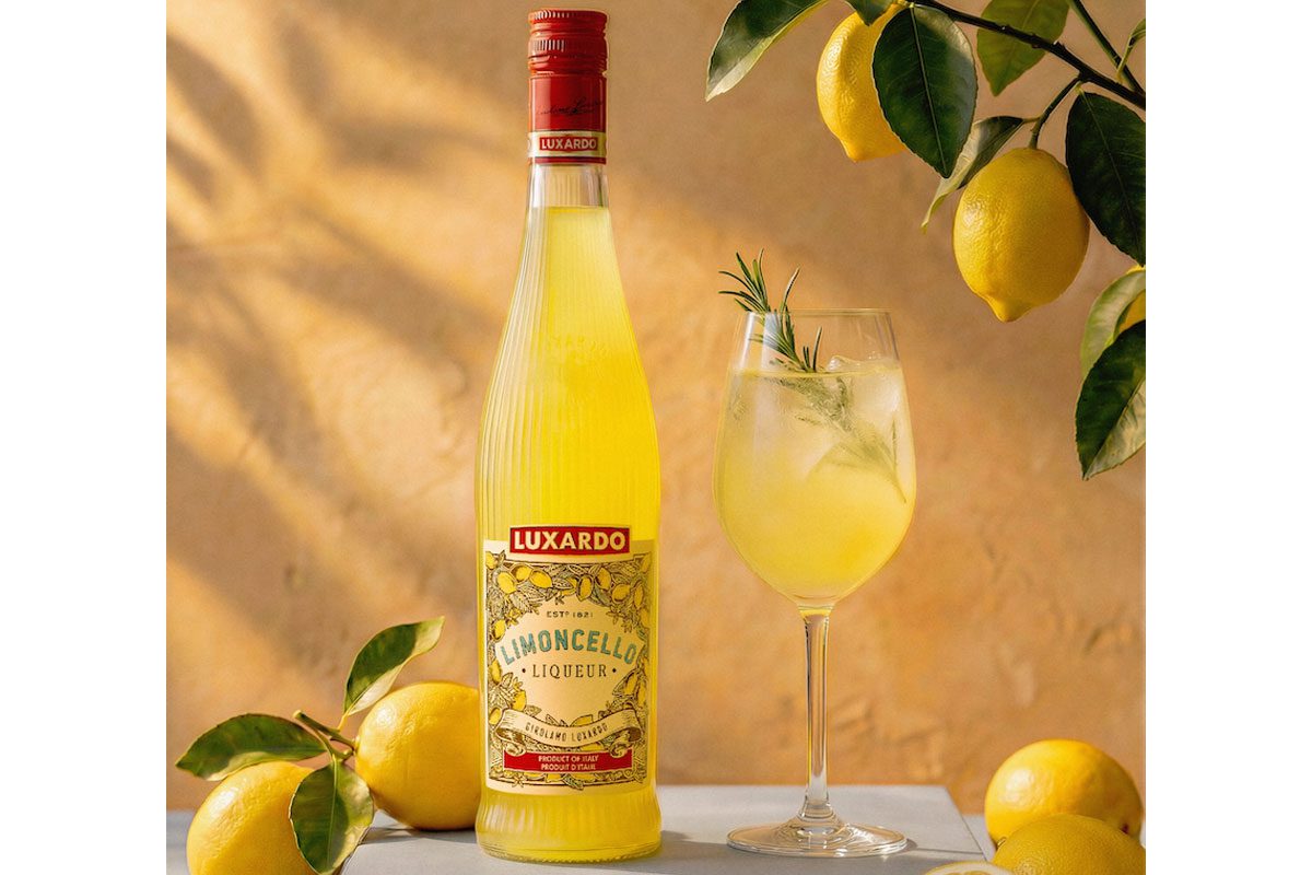 The 2026 redesign for Luxardo Limoncello stands on a table next to a wine glass with a cocktail serve in it and lemons have been placed around the two with a lemon tree hanging above them.