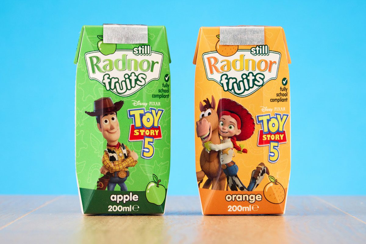 Packs of Radnor Hills Toy Story 5 collaboration in Apple and Orange flavours, these are set against a blue background on top of a wooden table.