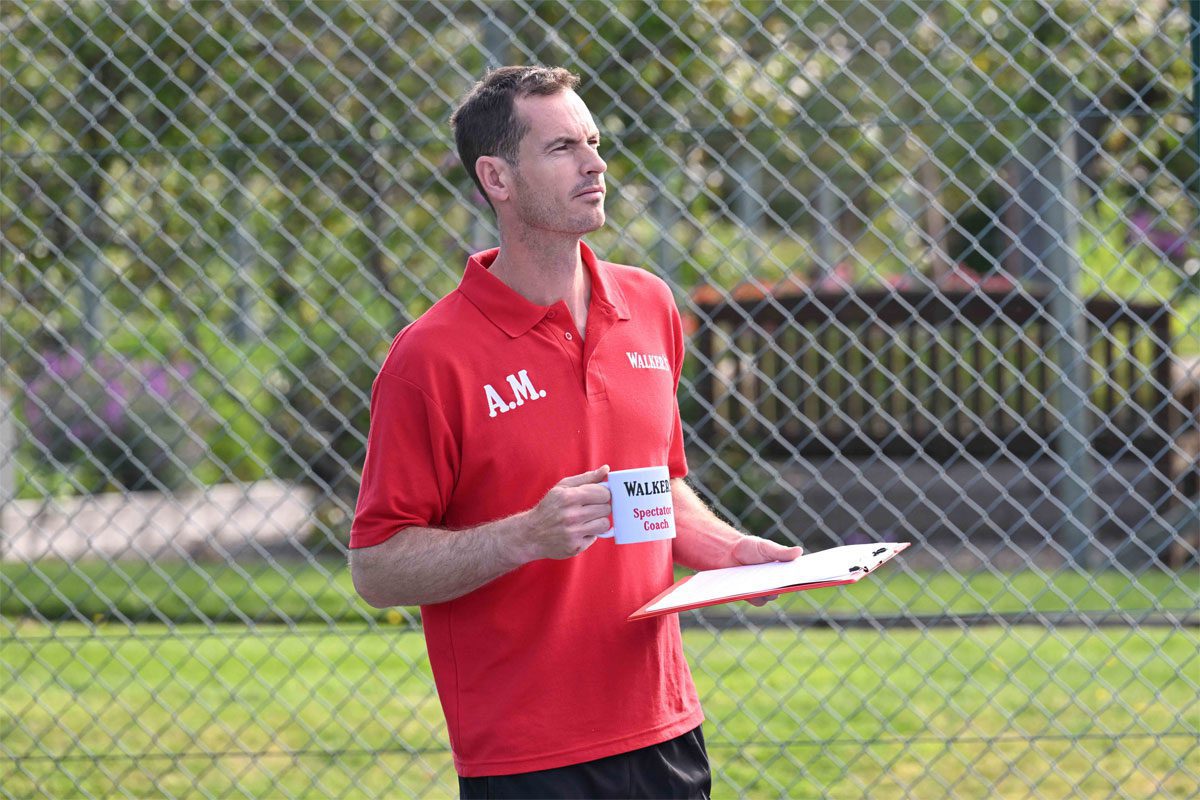 Andy Murray stands in a tennis court holding a mug that says 'Walkers Spectator Coach' on it.