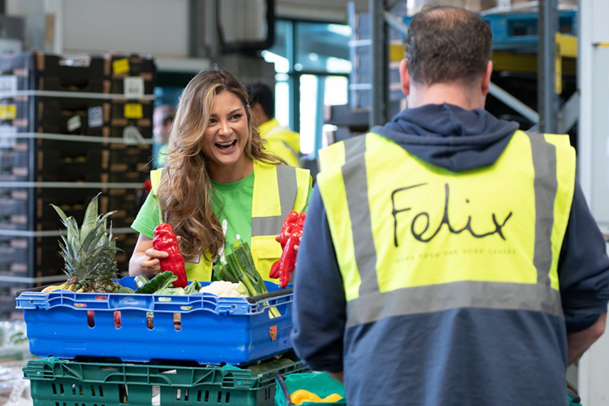 Two people in high-vis jackets stand talking whilst moving crates of surplus food. The jacket facing the camera features the Felix logo.