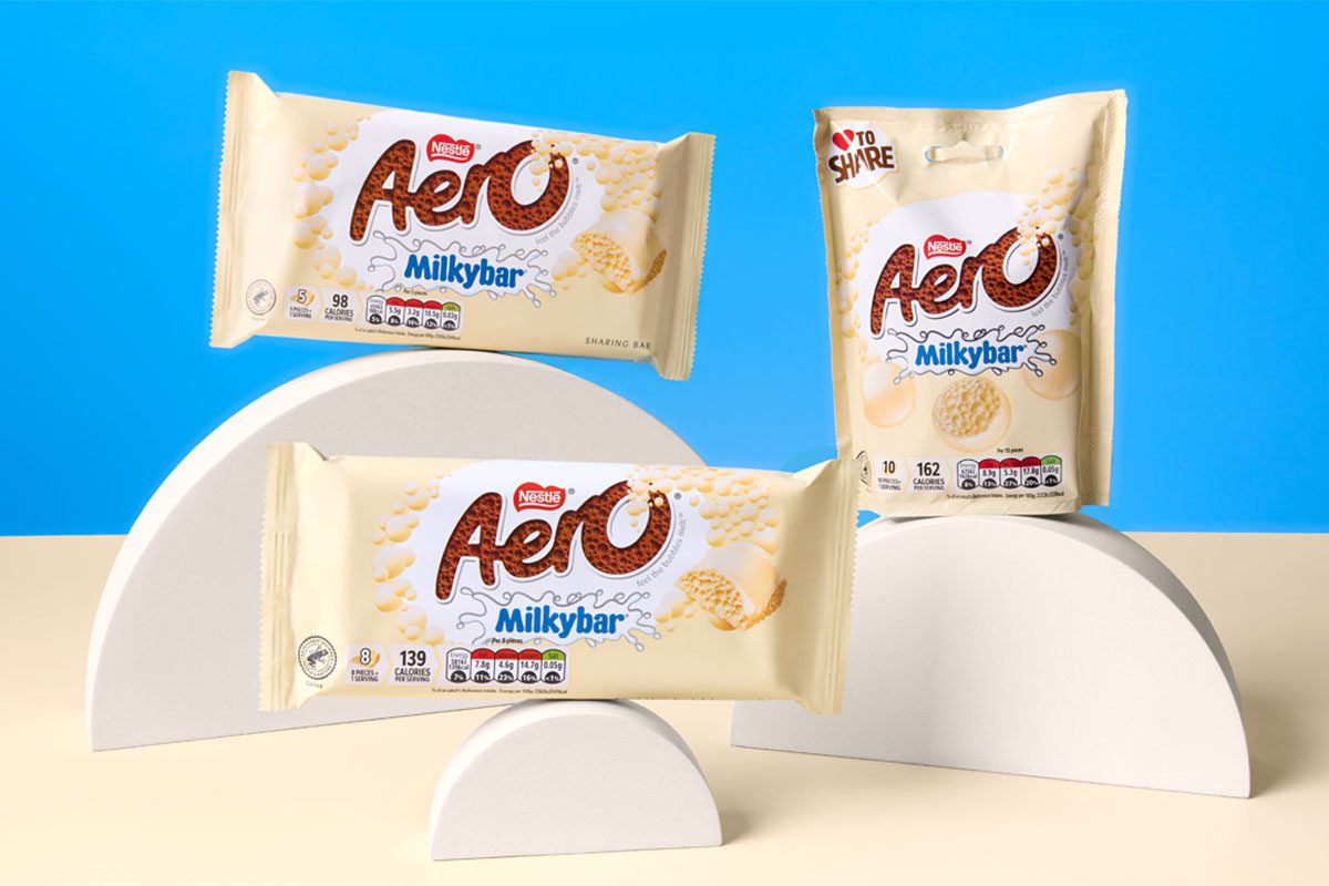 Packs of Aero Milkybar and Aero Milkybar Buttons are stacked on semi-circle shapes against a blue background with a white table.