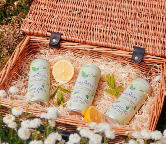 Darnley’s Gin enters the RTD market with Elderflower Spritz Cans of Darnley's Elderflower Gin Spritz lie in a basket with pieces of mint and lemon around them. The basket sits on a grassy field with flowers.