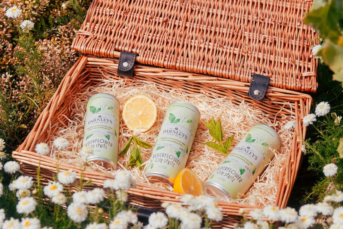 Cans of Darnley's Elderflower Gin Spritz lie in a basket with pieces of mint and lemon around them. The basket sits on a grassy field with flowers.