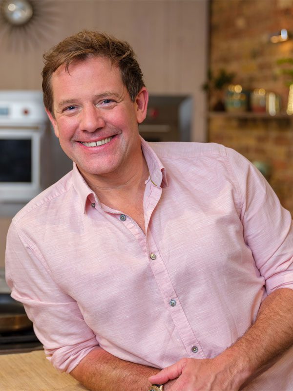 Matt Tebbutt stands in a kitchen leaning on the counter.