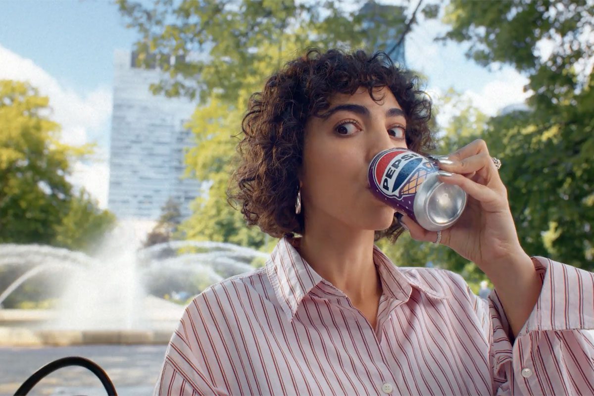 A woman sits drinking from a can of Pepsi Cherry & Vanilla Flavour in a park.