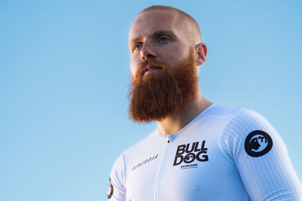 Endurance athlete Russ Cook stands against a blue background wearing a Bulldog Skincare branded top.