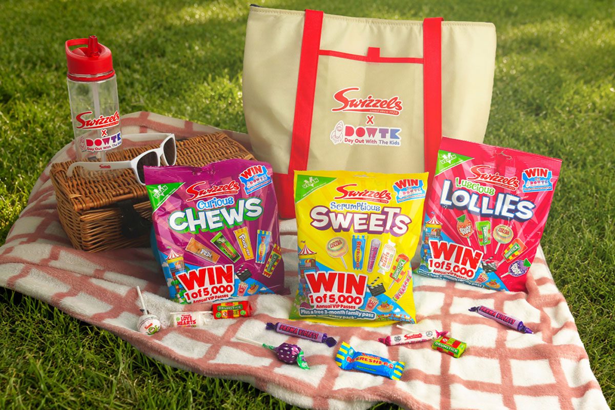 A picnic scene with variety packs of Swizzels sweets, a picnic blanket, a basket, sunglasses, a water bottle and an insulated bag with the Swizzels and Day Out With The Kids logo on the front of it.