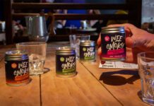 Wee Smoky taps into RTD demand with cocktail range A table with Wee Smoky cocktail cans on it and glasses with a person holding one of the cans in their hand.