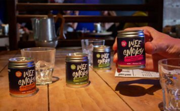 Wee Smoky taps into RTD demand with cocktail range A table with Wee Smoky cocktail cans on it and glasses with a person holding one of the cans in their hand.