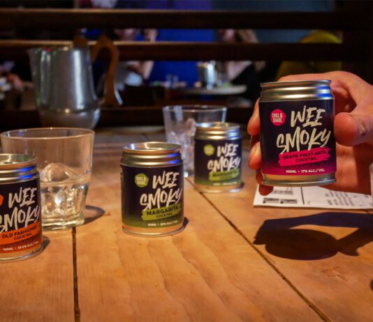 Wee Smoky taps into RTD demand with cocktail range A table with Wee Smoky cocktail cans on it and glasses with a person holding one of the cans in their hand.