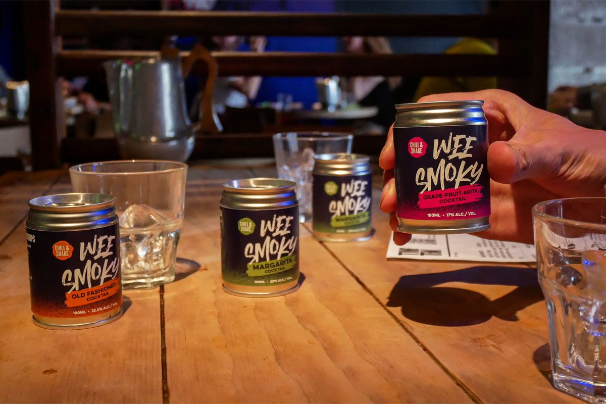 A table with Wee Smoky cocktail cans on it and glasses with a person holding one of the cans in their hand.