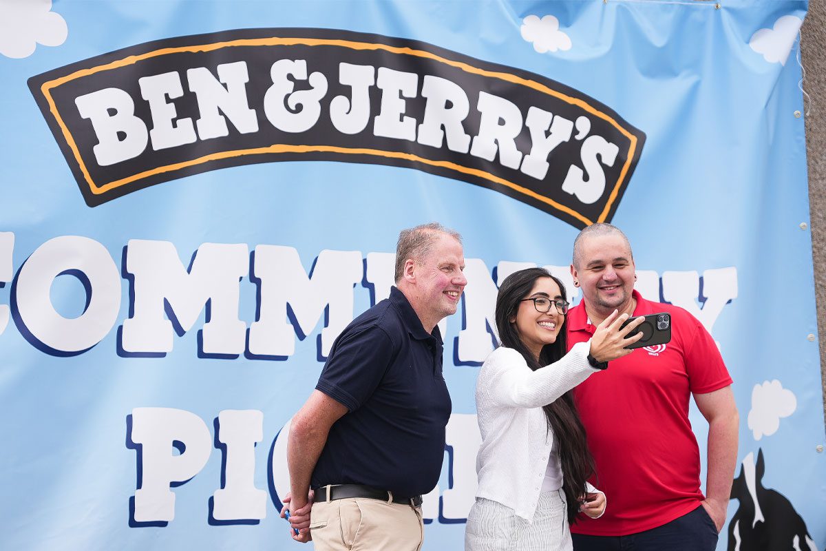Chris Cobb, store owner of Cults Stores – KeyStore, stands with Ghayda Jameel, senior account exec at Cirkle, and Andrew Young, business development exec at The Magnum Ice Cream Company, stand together to take a selfie in front of the Ben & Jerry's Community Picnic banner. 