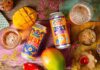 Curry Smugglers launches new Desi Beer brand Cans of Desi Lager Basmati Rice Lager and alcohol-free Mango Mirchi Lager lay on a table with food ingredients around it and glasses with the beers poured into them.