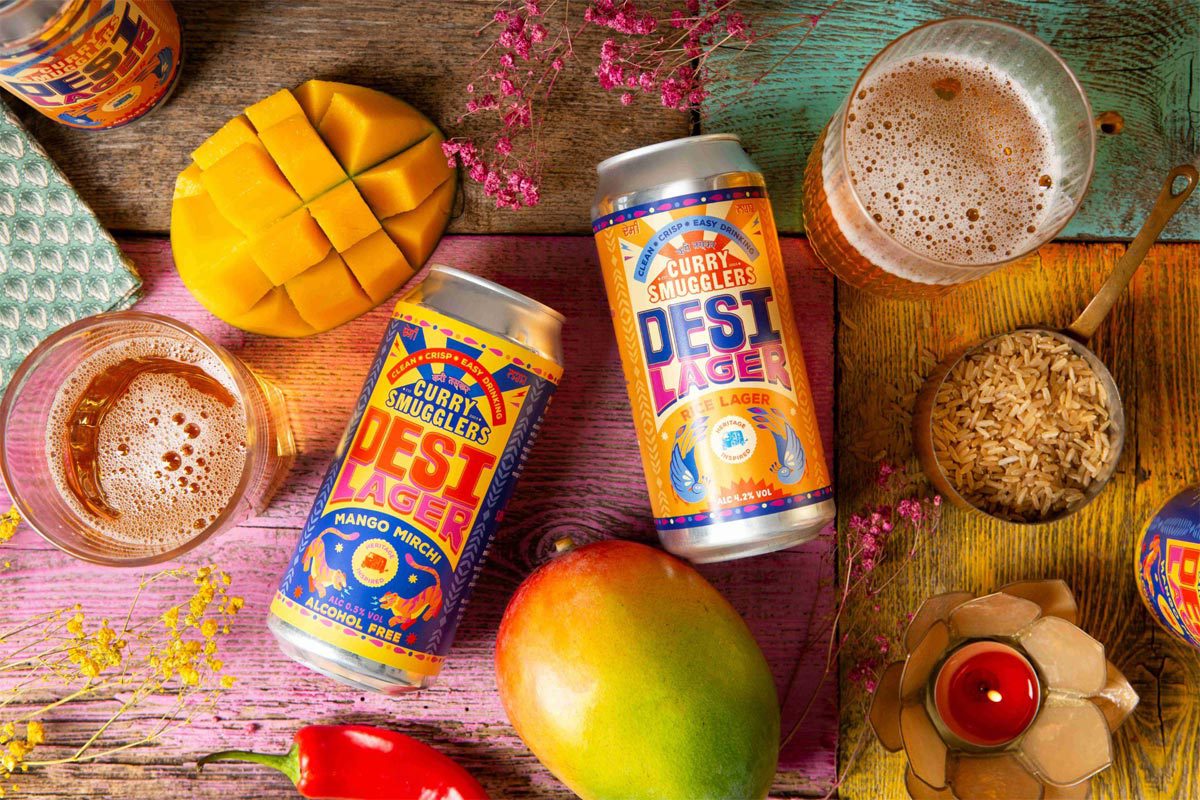 Cans of Desi Lager Basmati Rice Lager and alcohol-free Mango Mirchi Lager lay on a table with food ingredients around it and glasses with the beers poured into them.
