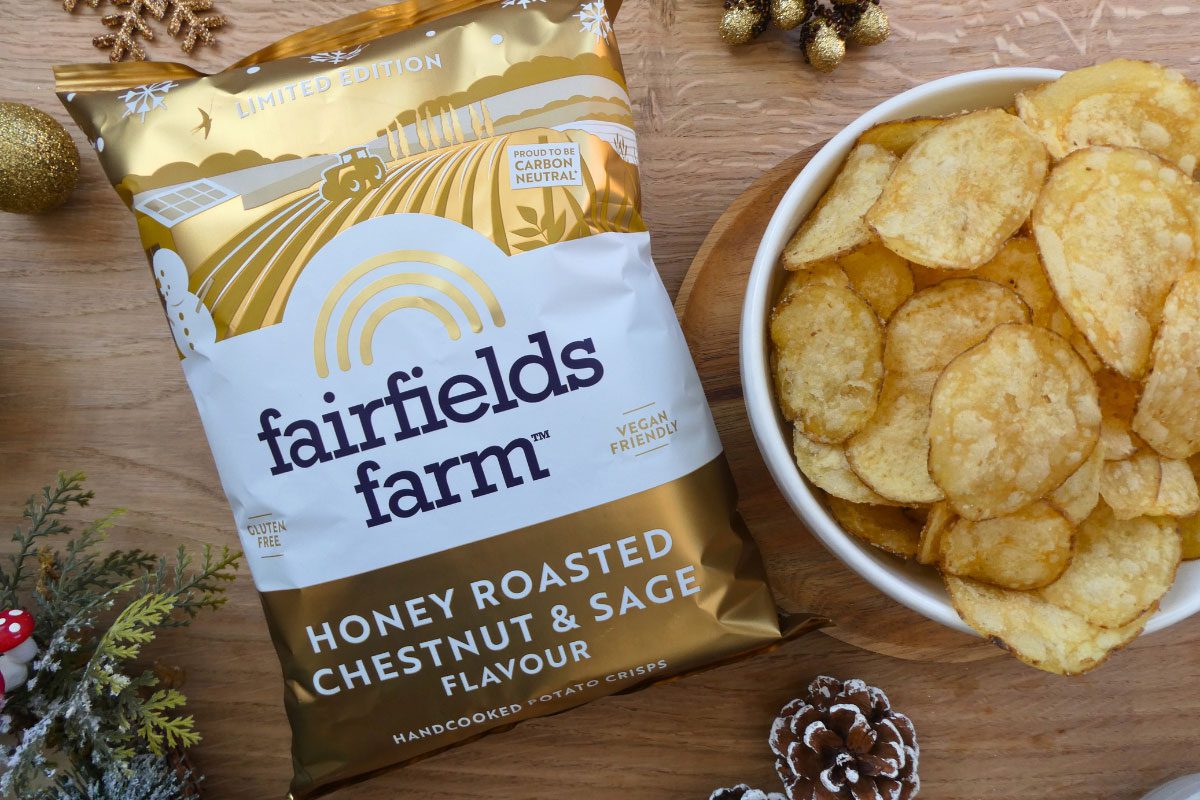 A pack of Fairfields Farm Honey Roasted Chestnut & Sage crisps sits next to a bowl with crisps in it with some Christmas decorations around the two.