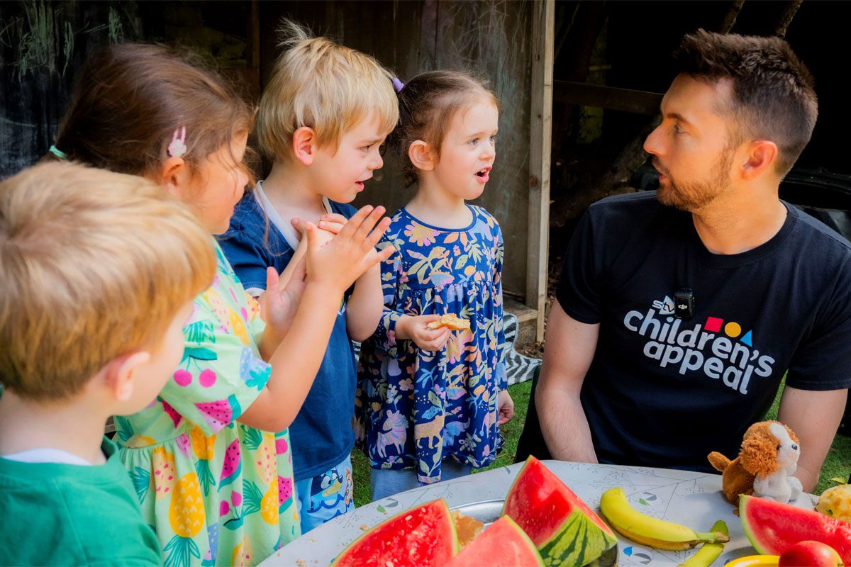 STV presenter Sean Batty sits at a table with fruit on it talking to a group of children.