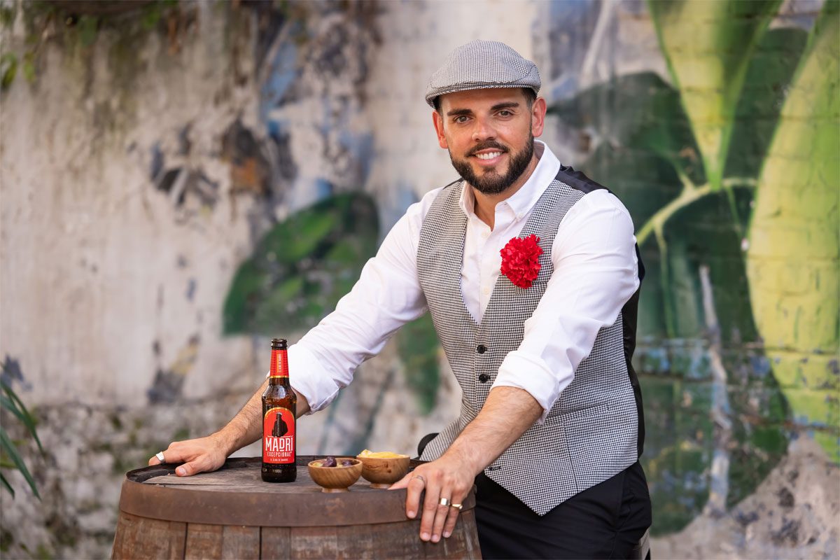 A man dressed like the mascot of MadrĂ Excepcional stands at a barrel in a street with a bottle of the beer on top of the barrel.